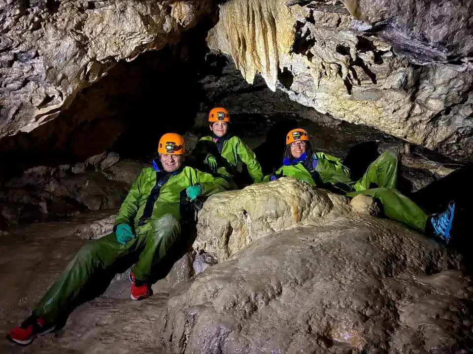 Trois personnes souriantes, équipées de casques et de combinaisons de spéléologie, assises dans une grotte du Dévoluy qui est ornée de stalactites et de formations rocheuses humides.
