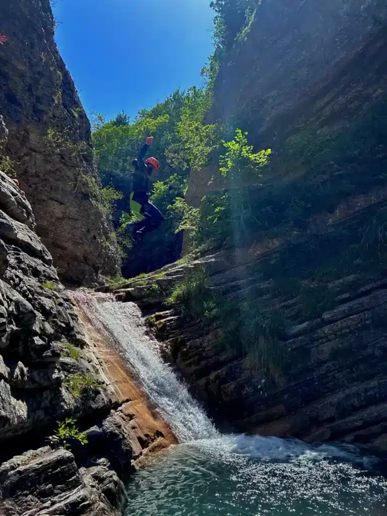Personne en combinaison de canyoning et casque orange saute d'une falaise dans un bassin d'eau au pied d'une petite cascade dans le canyon de Rabou..