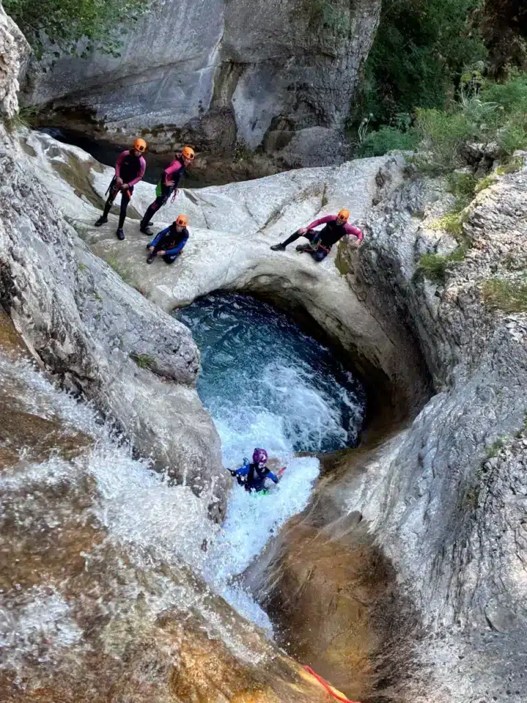 Un groupe de personnes en tenue de canyoning avec des casques de couleur orange admirent et effectuent un saut dans une cascade d'eau cristalline creusée dans la roche du canyon de Rabou.
