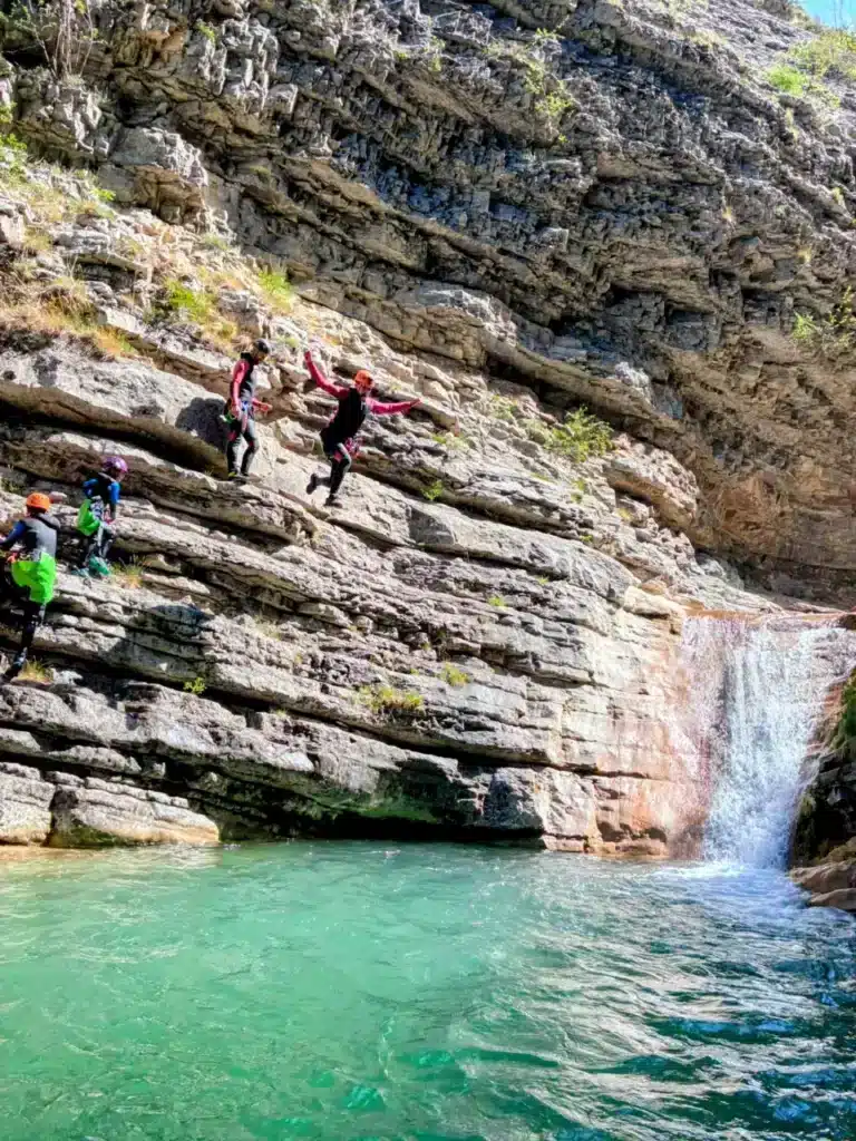 Des personnes en tenue de canyoning s'élancent d'une falaise rocheuse au-dessus d'une piscine naturelle d'eau turquoise bordée par la petite cascade du canyon de Rabou.