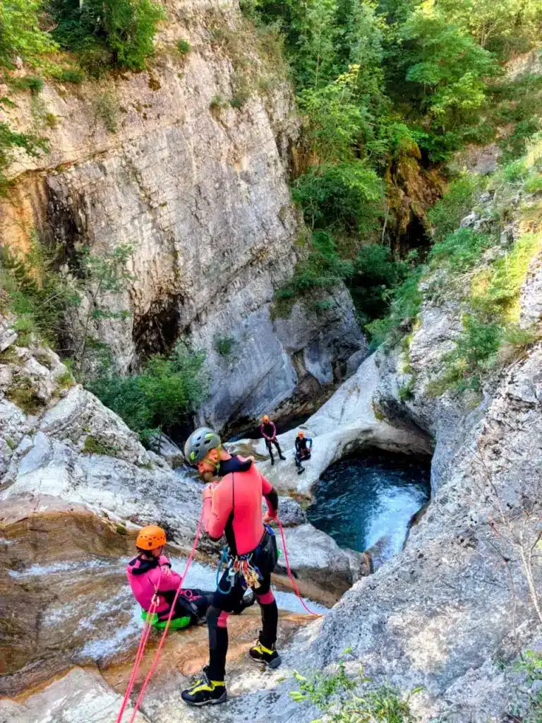 Trois personnes en combinaison de canyoning descendent en rappel dans le canyon de Rabou, en contrebas un bassin d'eau bleue.