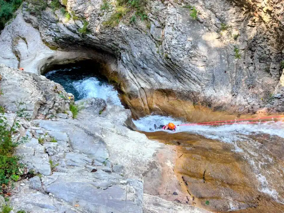 Une personne en équipement de canyoning orange descend une cascade dans le canyon de Rabou aux parois rocheuses abruptes, tenue par une corde rouge.