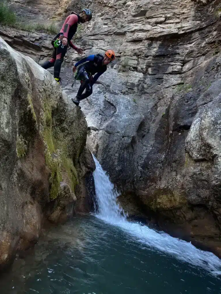 Deux personnes en combinaison de canyoning s'apprêtent à sauter dans une vasque d'eau du canyon de Rabou, une vue près de l'eau turquoise.