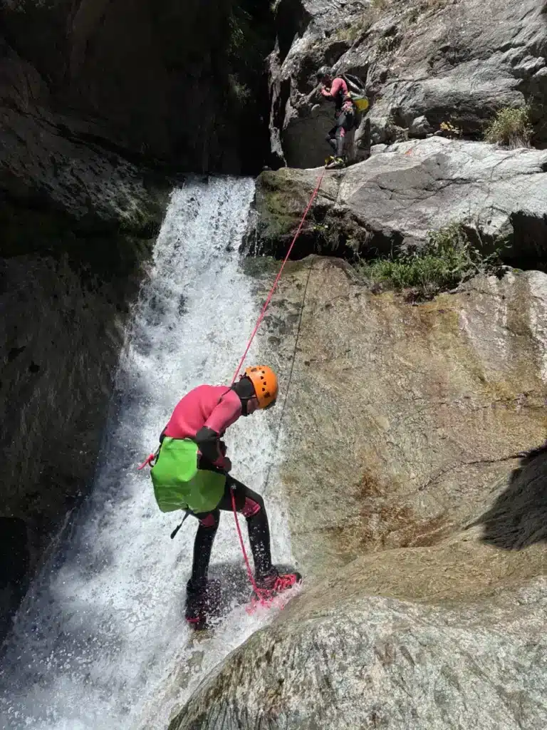 Deux personnes en combinaison font du canyoning dans une rivière à Orcières, l'une descendant en rappel une cascade, l'autre observant depuis le haut.