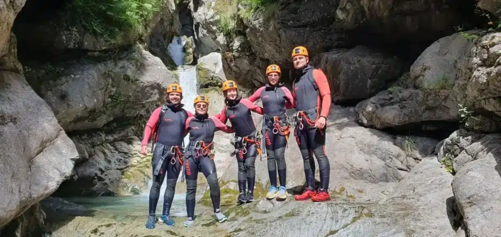Groupe de cinq personnes en combinaison de canyoning et casques, souriant dans le canyon de Val Estrèche à Orcières Merlette avec une petite cascade derrière eux.