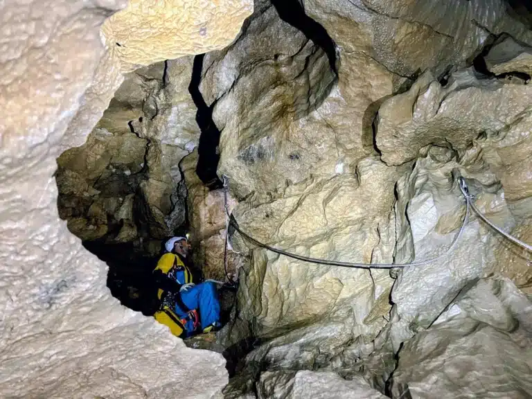 Spéléologue équipé d'un baudrier et d'un casque, assis dans une grotte, à côté d'une paroi de la viasouterrata de SuperDévoluy