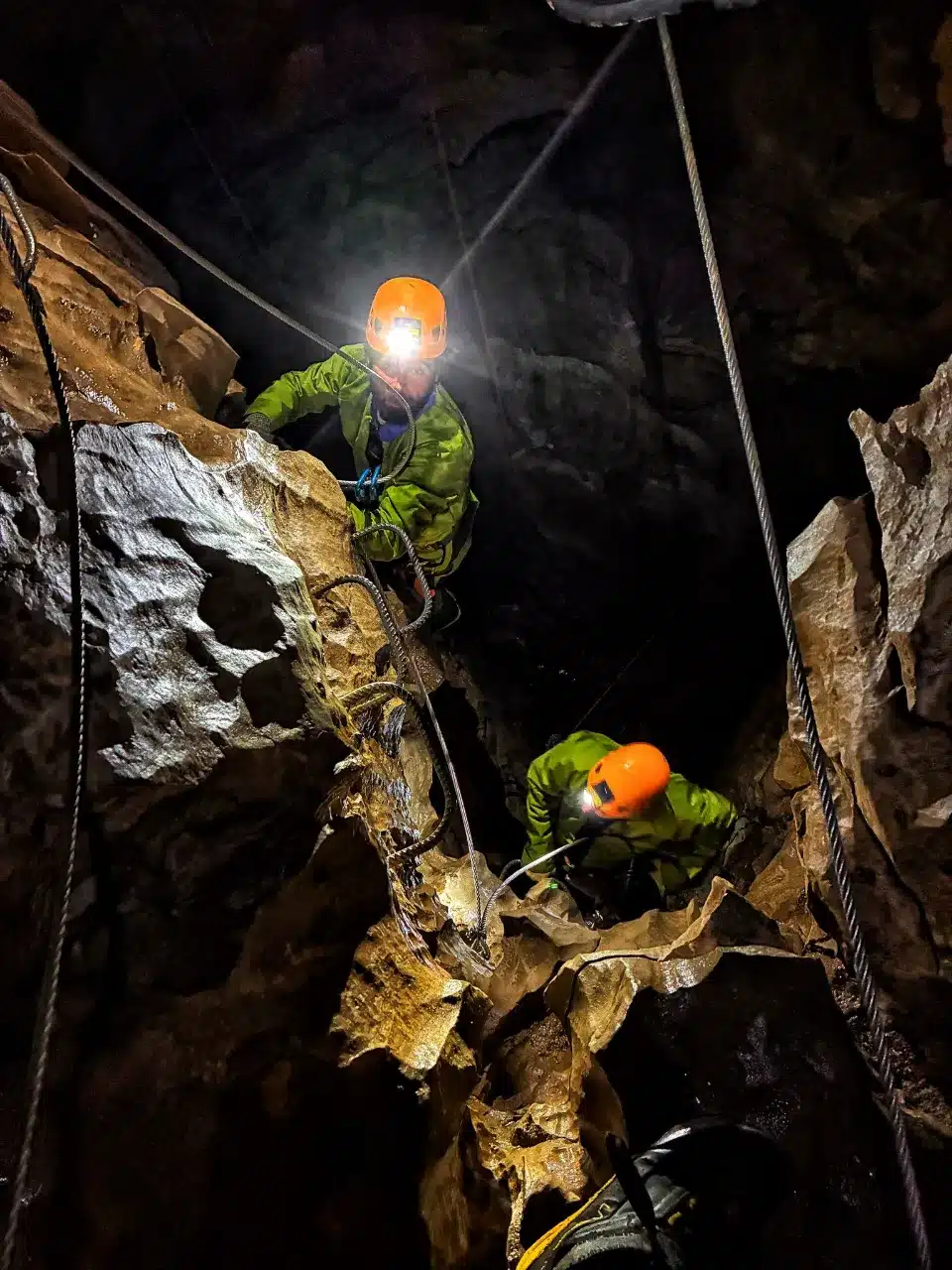 Deux spéléologues équipés de casques lumineux et de combinaisons vertes descendent dans la via souterrata de SuperDévoluy, assurés par des cordes.
